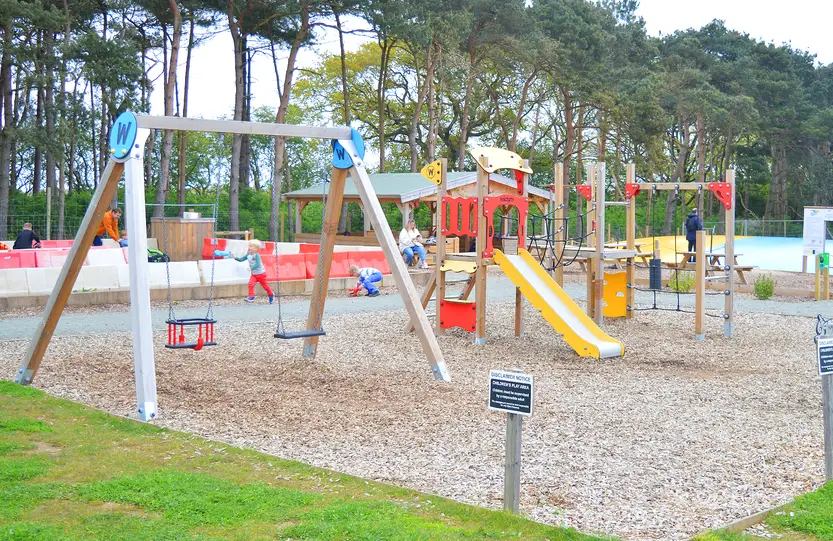 Outdoor children's play area at The Bug Parc featuring swings, climbing equipment and slides set among mature woodland.