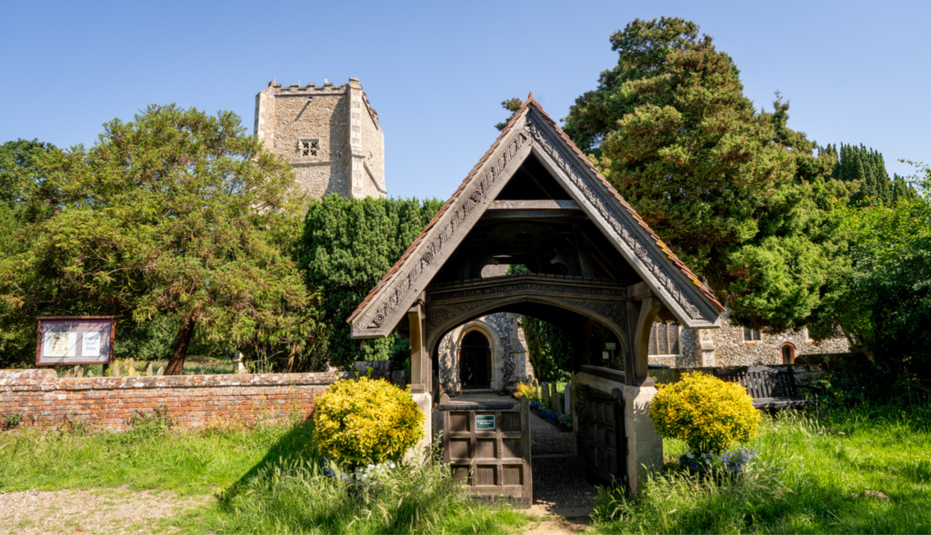 Traditional timber-framed lychgate at St Mary's Church, Salhouse, with the historic church tower visible behind and surrounded by mature trees and landscaped churchyard gardens.