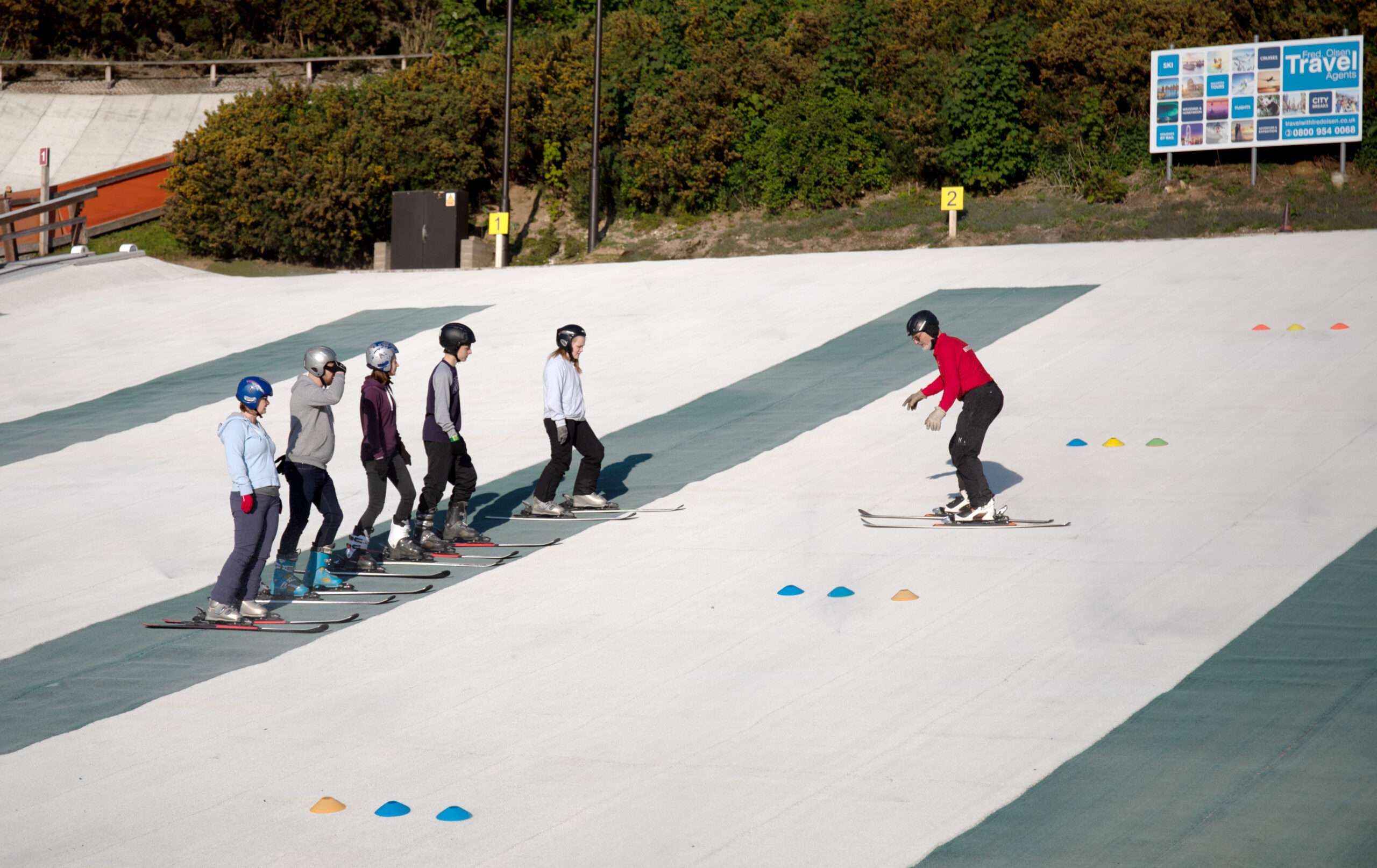 Group ski lesson on a gentle artificial ski slope, with participants following an instructor as another skier descends nearby.