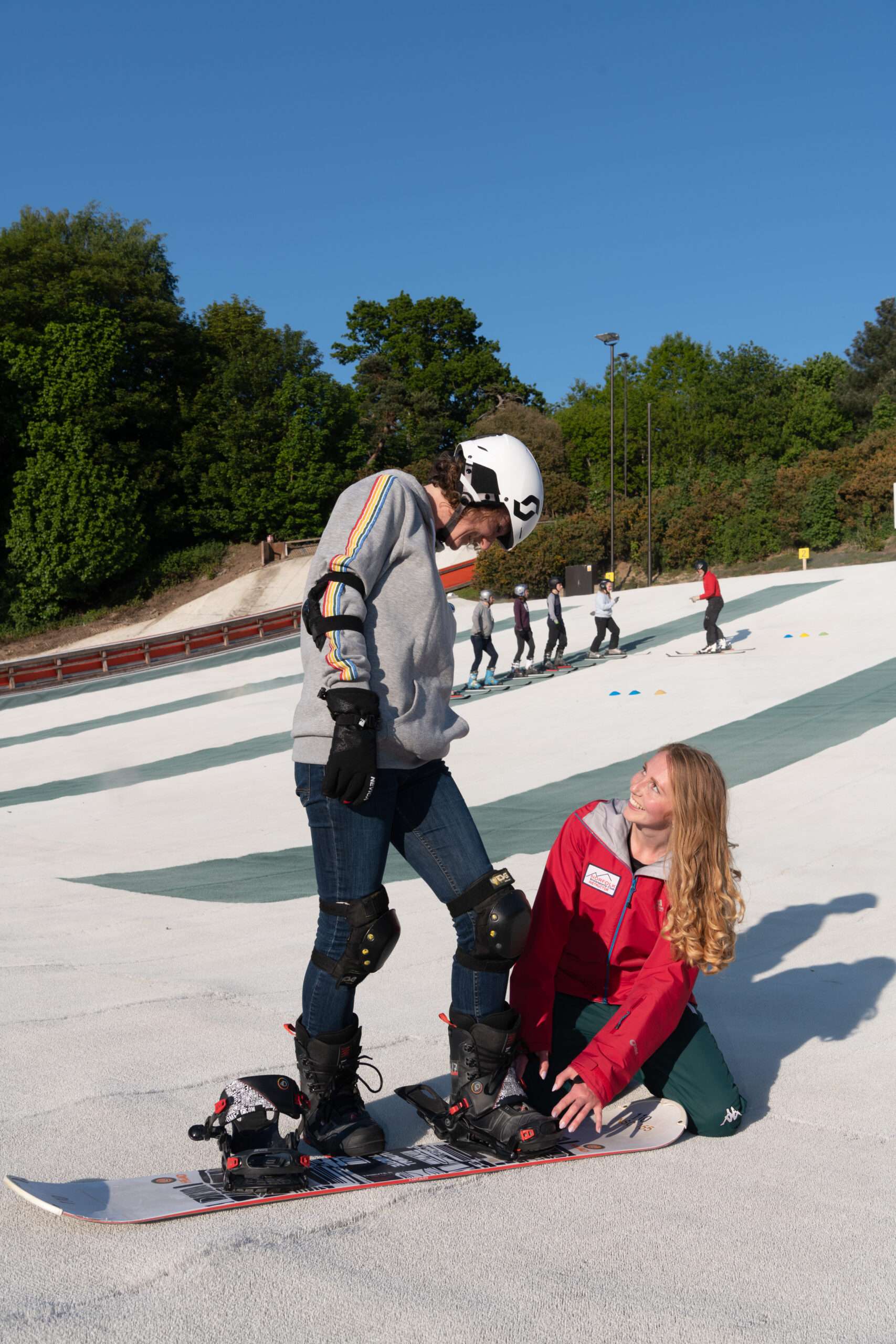 Person in winter clothing speaking with a seated woman on a beginner ski slope, with snow-covered practice areas and trees in the background.