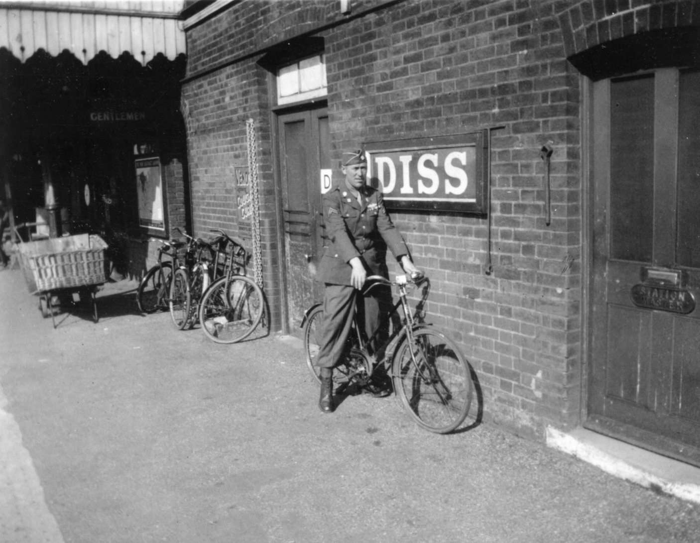Historic photograph of soldier on a bicycle outside Diss railway station platform and station signage.
