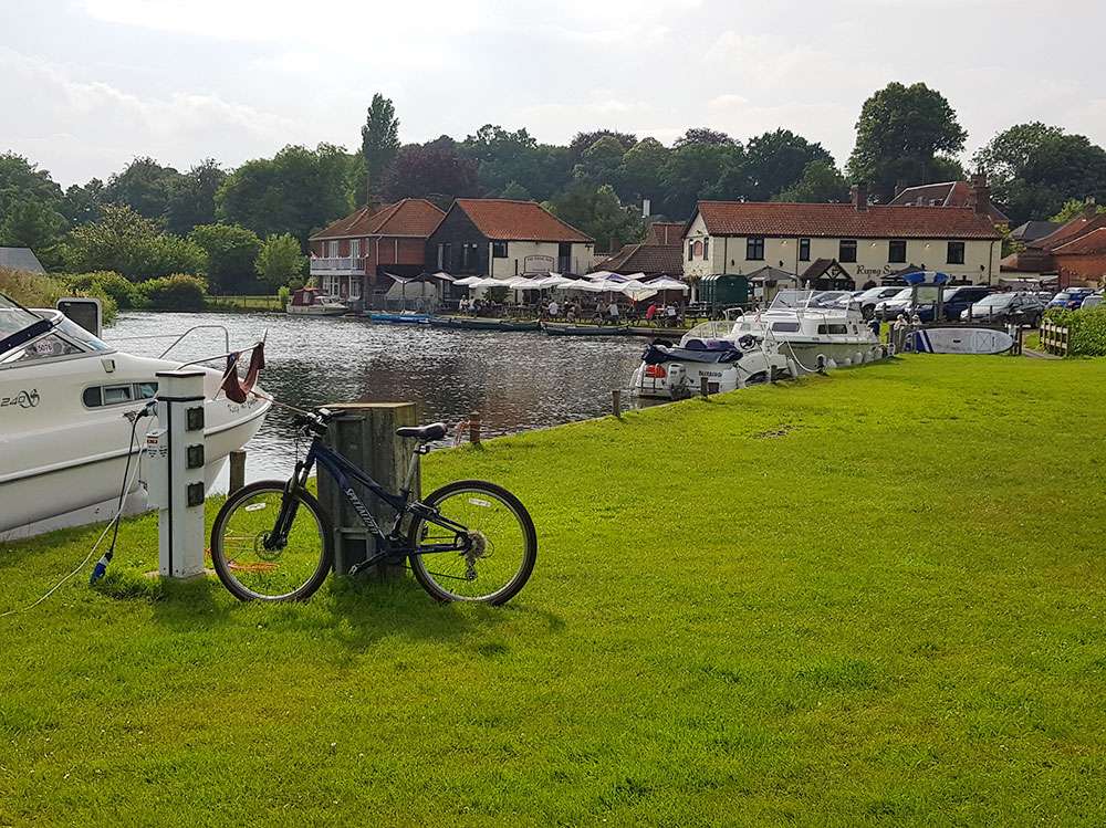 View of Bure Valley Cycle Hire beside the River Bure, with boats moored along the waterfront and Norfolk countryside in the background.