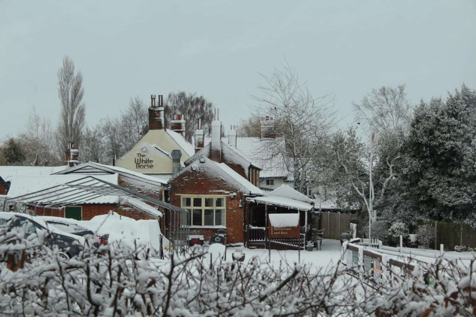 White Horse pub in Loddon during winter snowfall, with snow-covered rooftops, trees and outdoor seating beside the traditional Norfolk pub.