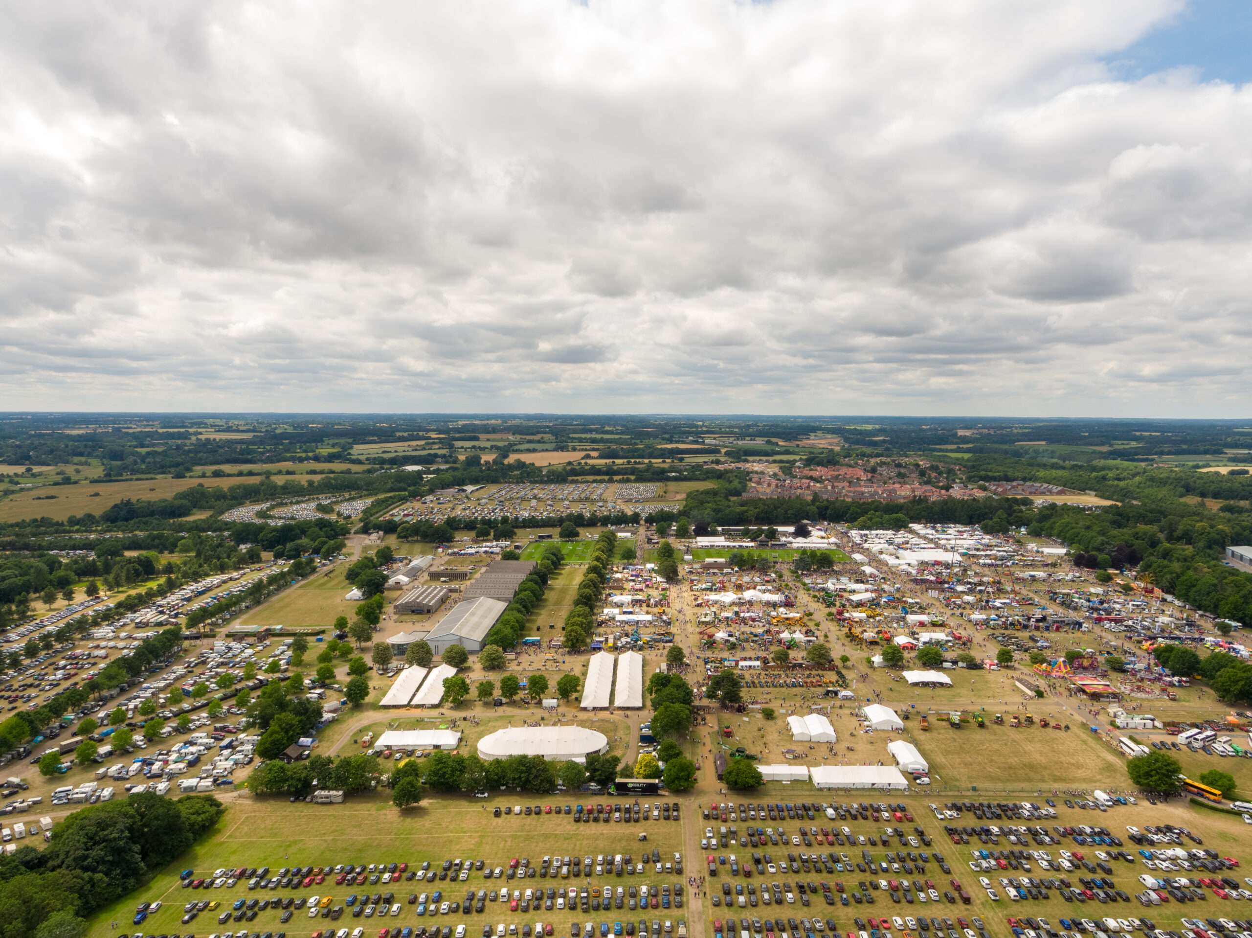 Aerial view of the Royal Norfolk Showground with exhibition areas, marquees, attractions, car parks and surrounding countryside under a cloudy sky.