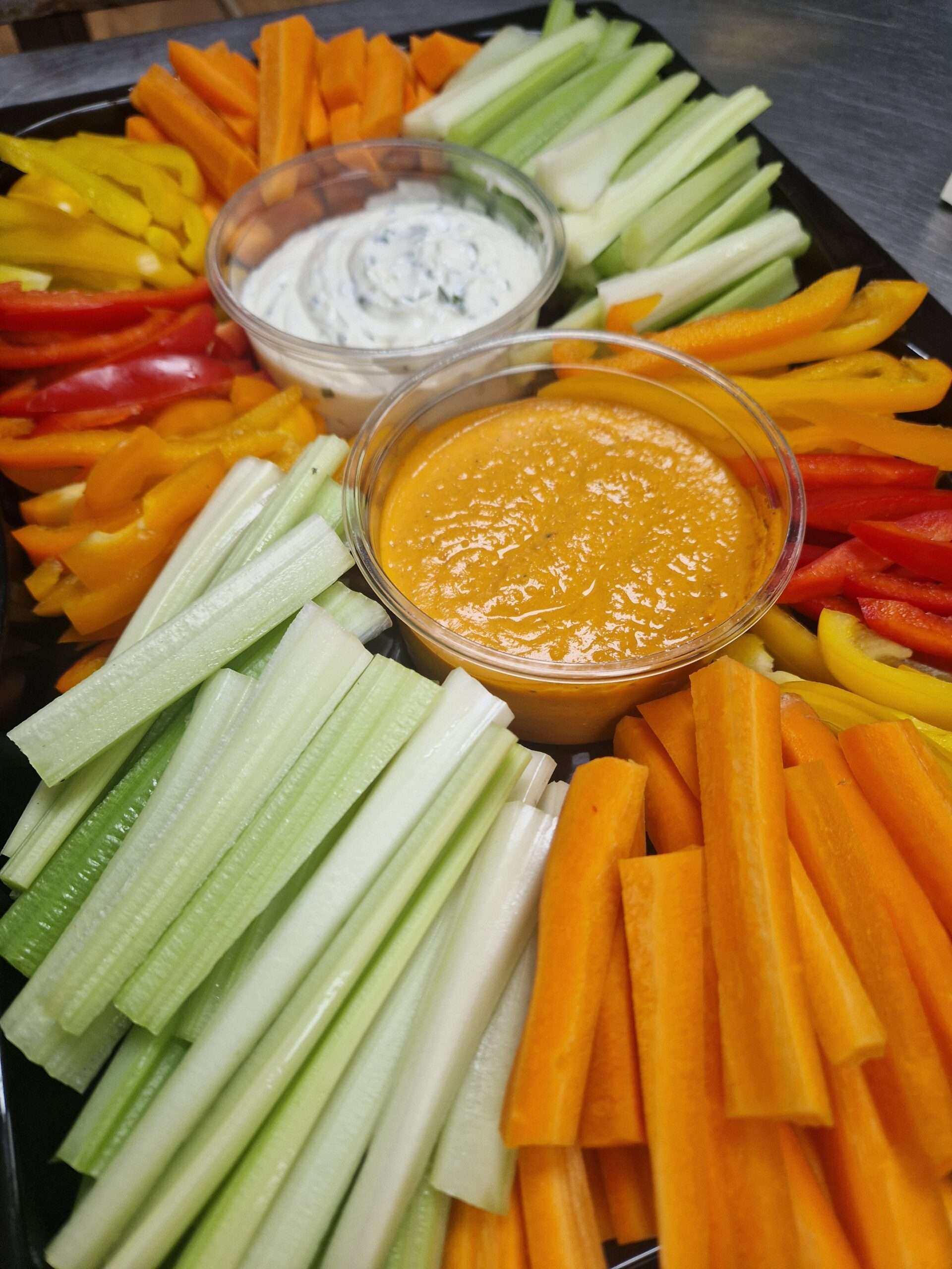Fresh vegetable crudités including carrot, celery and pepper sticks arranged around bowls of creamy dip and orange hummus.
