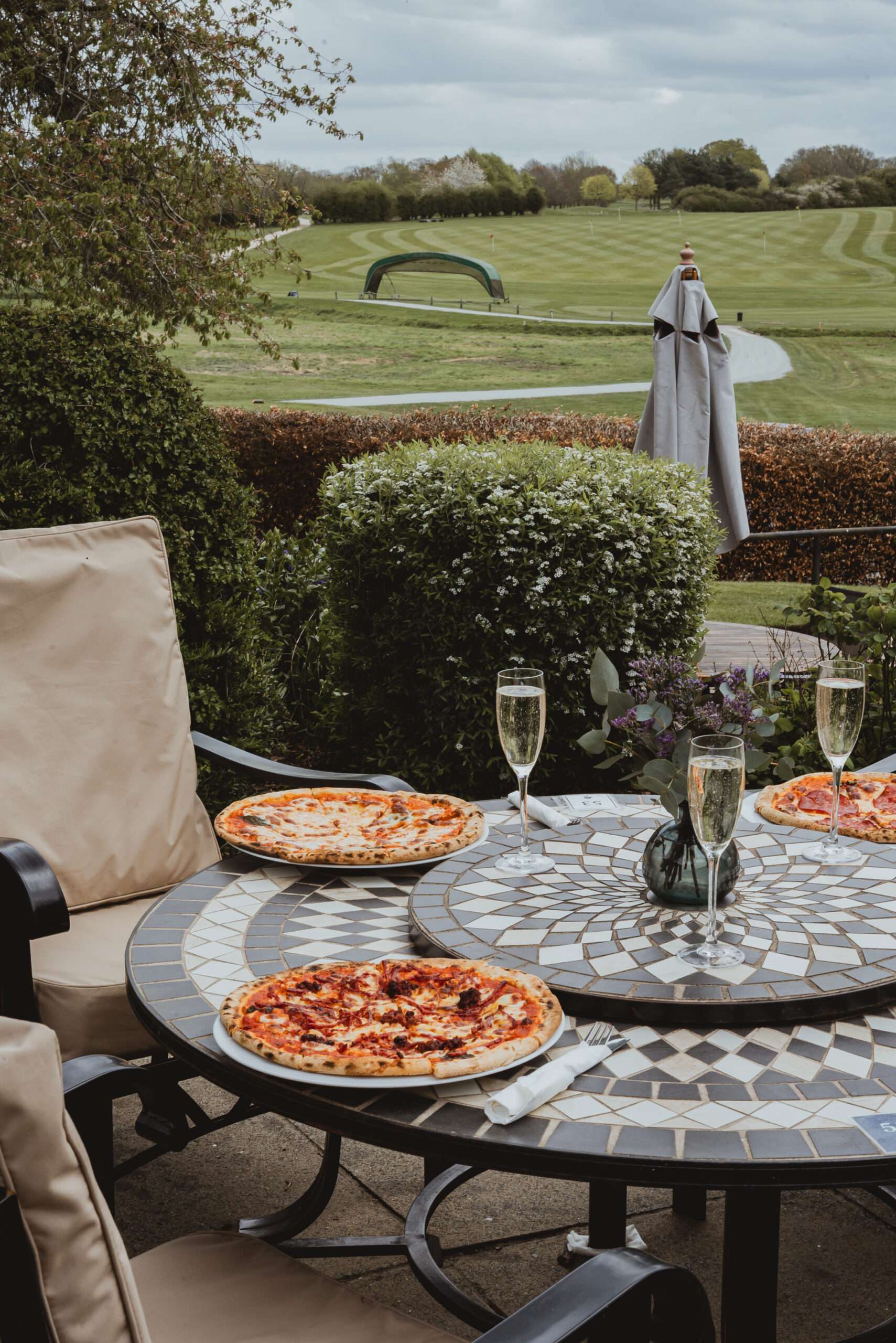 Outdoor dining table with pizza and prosecco overlooking landscaped gardens and countryside views at Barnham Broom Hotel, golf, and spa.