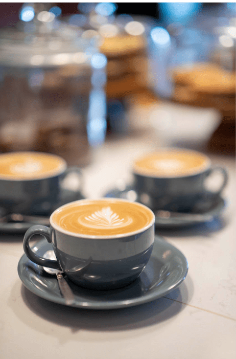 Latte art coffee served on a café table with additional cups and a blurred café background at Herbie's Farm Café, Salle Moor.