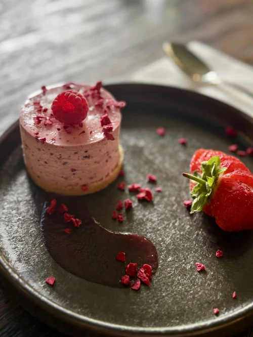 Dessert served on a dark plate, topped with fresh raspberries and scattered berry pieces, presented as part of a dining experience at The Huntsman, Strumpshaw.